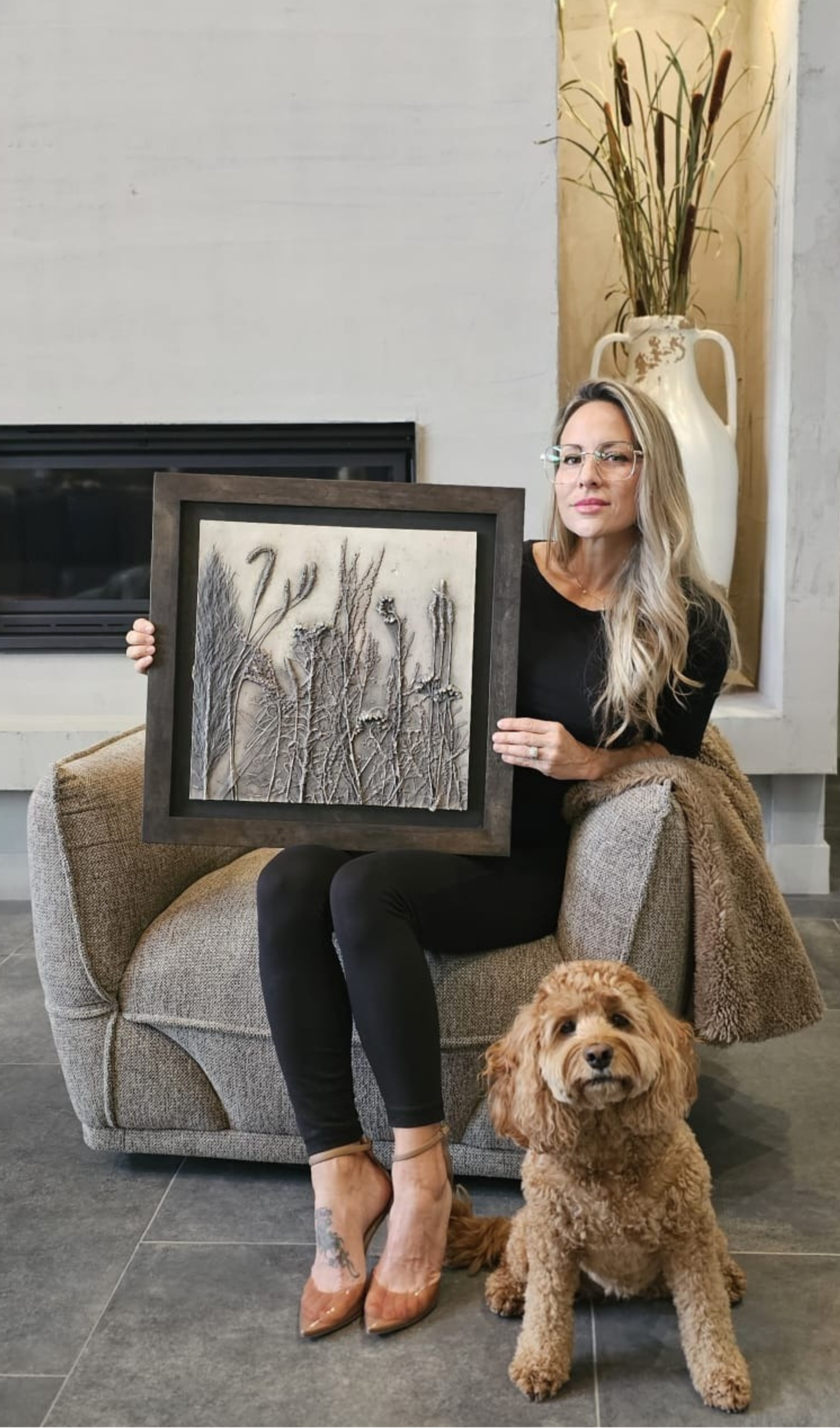 Une femme assise sur un fauteuil beige, tenant un tableau représentant une scène de nature avec des branches. Elle porte des lunettes, un vêtement noir et des talons beiges, avec une chatte posée devant elle. Un chien caniche marron clair est également devant elle, assis sur le sol.