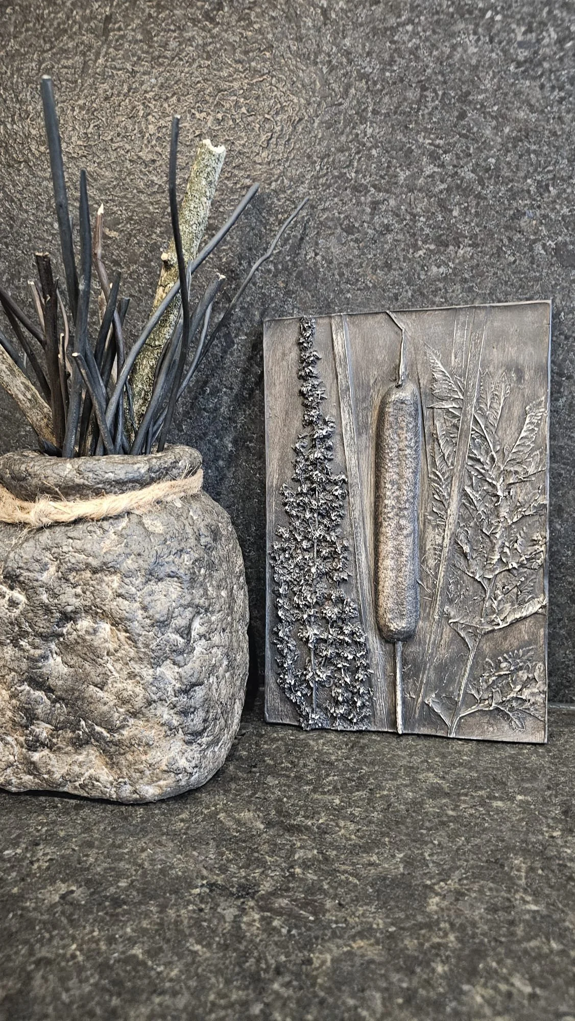 Un pot en pierre contenant des branches fines et sèches. En arrière-plan, une œuvre en relief représentant des plantes séchées en métal ou en bois.