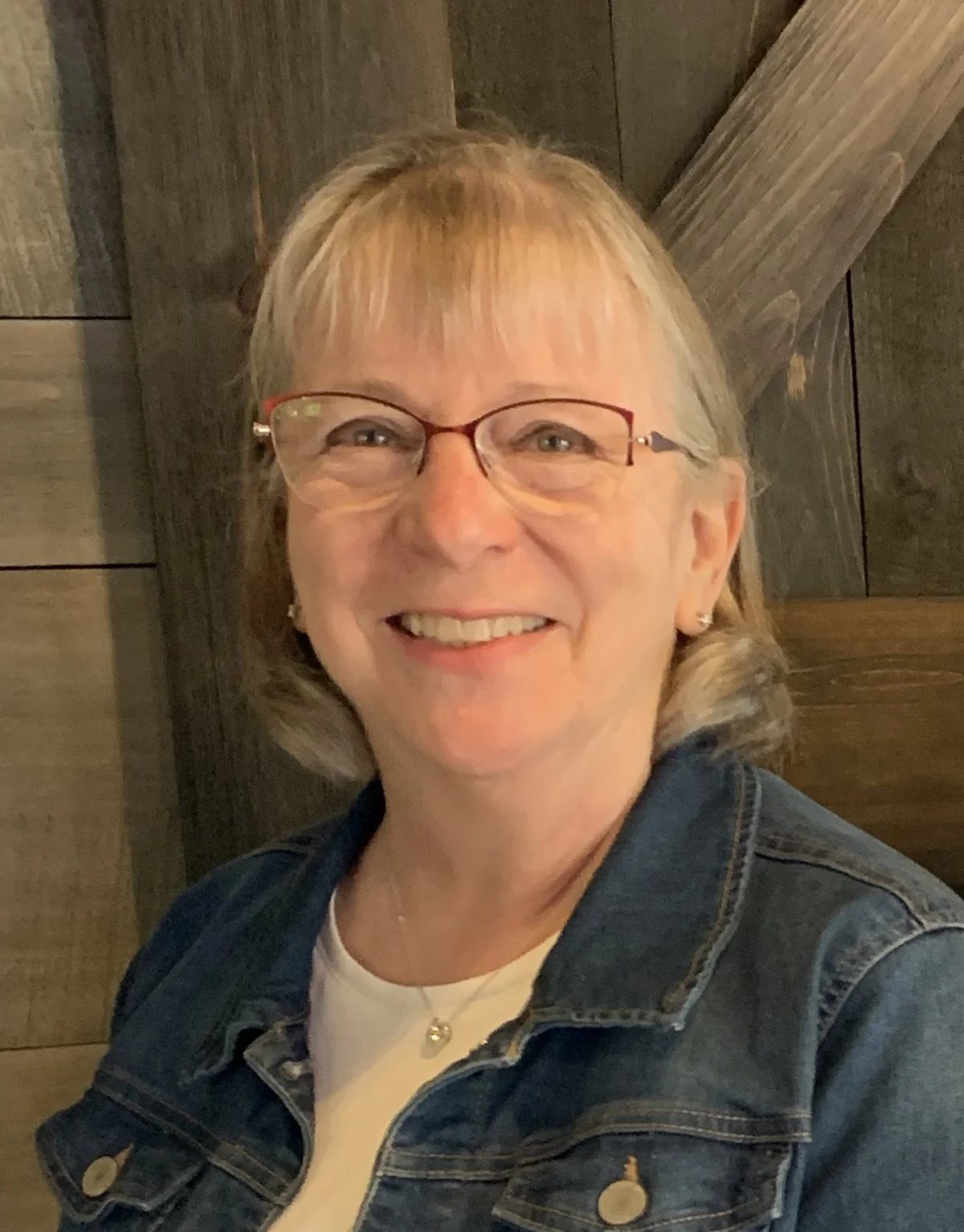 Smiling woman with glasses, short blond hair, wearing a denim jacket and jewelry, standing against a wooden background.