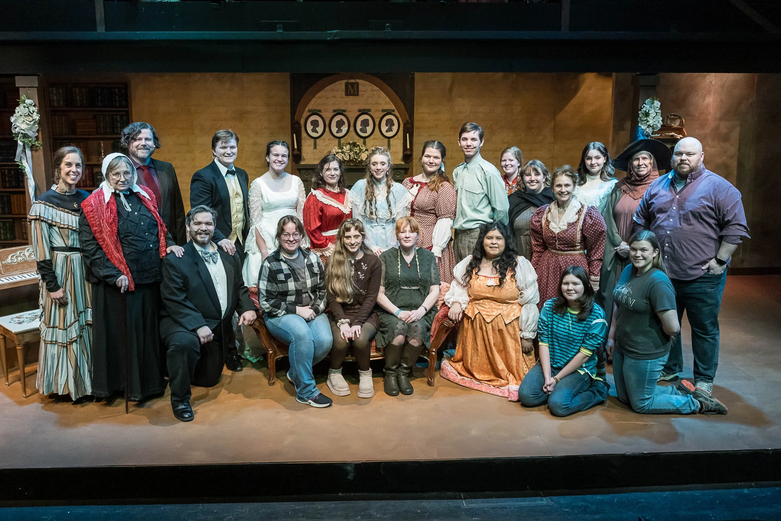 A group of people in historical costumes posing for a photo in a theater setting with a bookshelf and decorative items in the background.