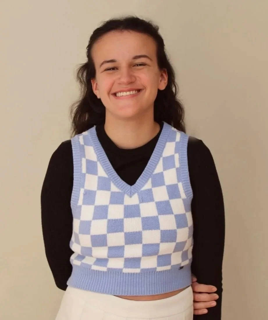 A young woman with dark brown, curly hair styled back, smiling and wearing a black long-sleeve shirt under a blue and white checkered sleeveless sweater vest, standing against a plain beige wall.