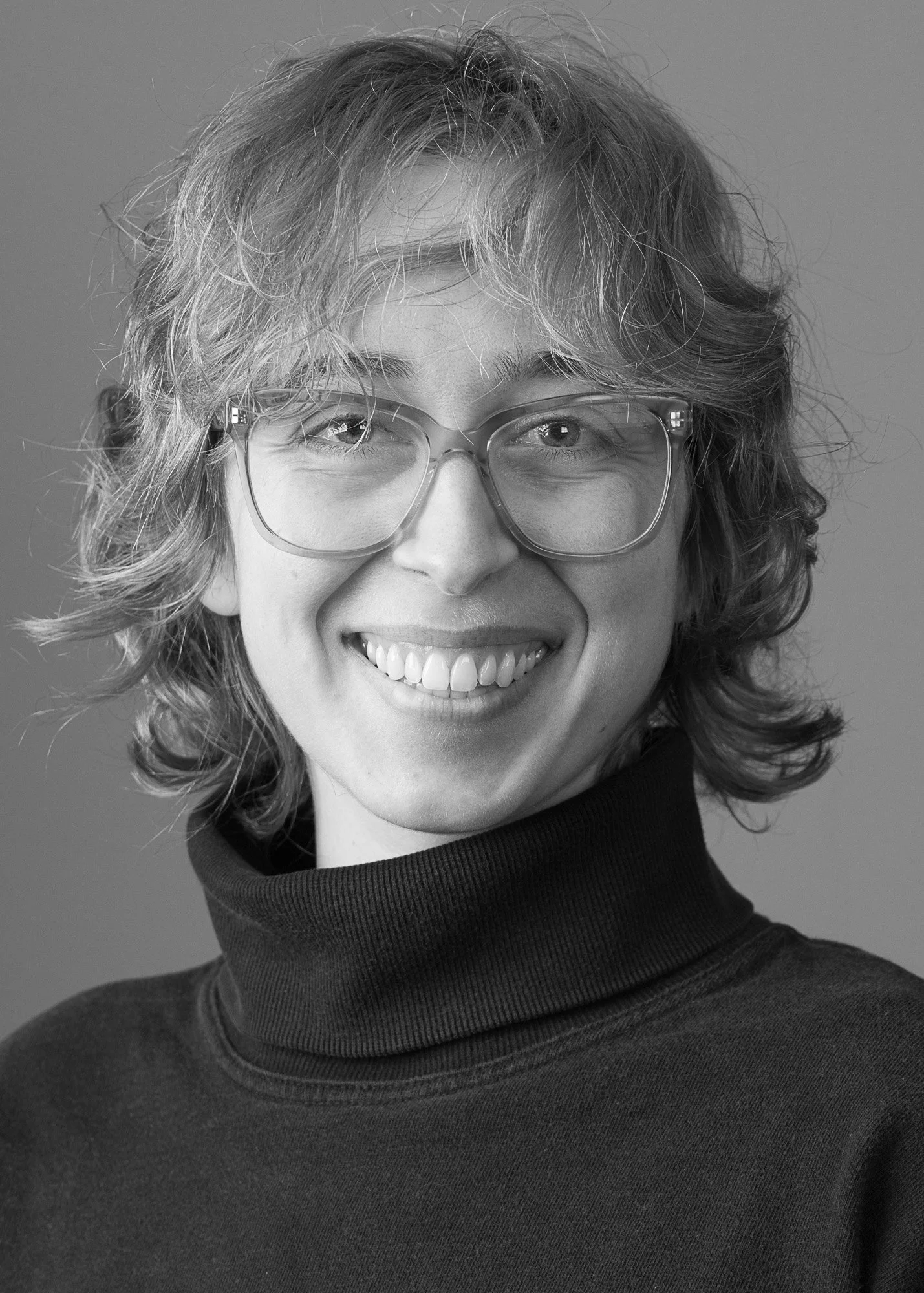 Black and white portrait of a smiling woman with curly hair, wearing glasses and a turtleneck sweater.