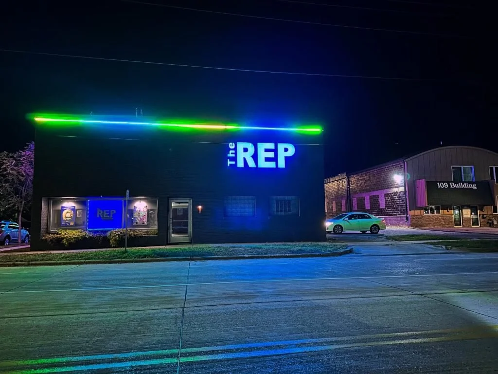 Nighttime exterior view of a building with a large illuminated sign that reads 'The REP' in blue lights. The building has a black facade with neon green and blue lighting accents. There are a few cars parked nearby, and adjacent buildings are visible in the background.