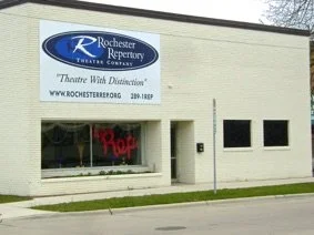 Exterior of Rochester Repertory Theatre building with a sign that reads 'Theatre With Distinction' and website URL, with a window showing a red neon sign inside.