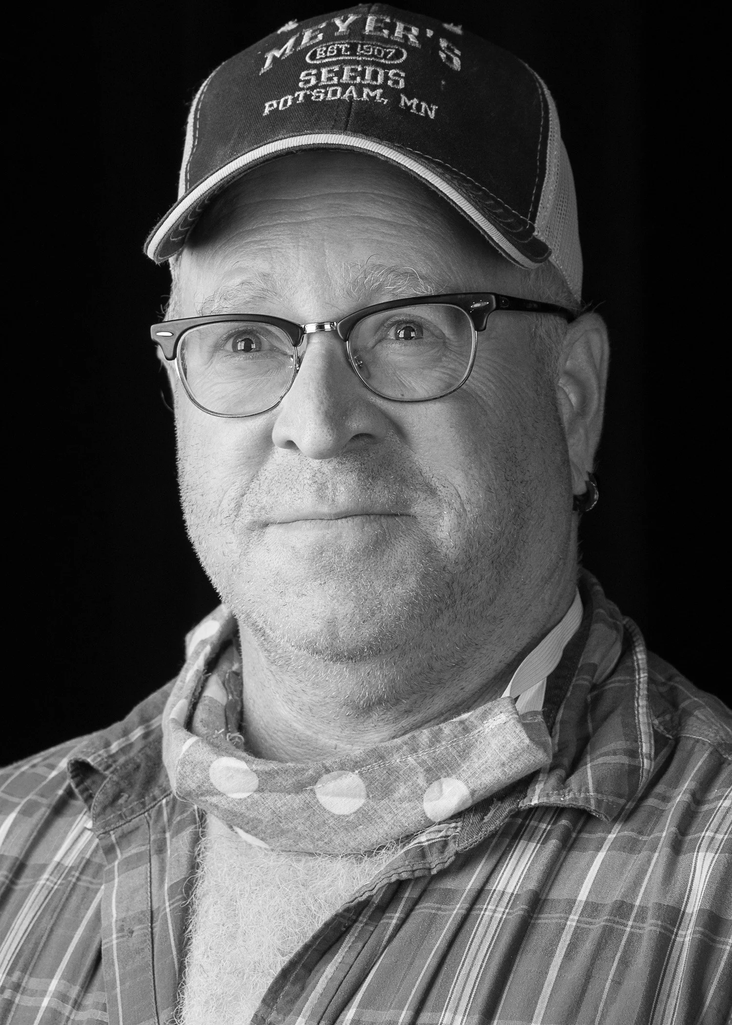 Black and white photo of a middle-aged man wearing glasses, a plaid shirt, a scarf around his neck, and a baseball cap with writing on it.