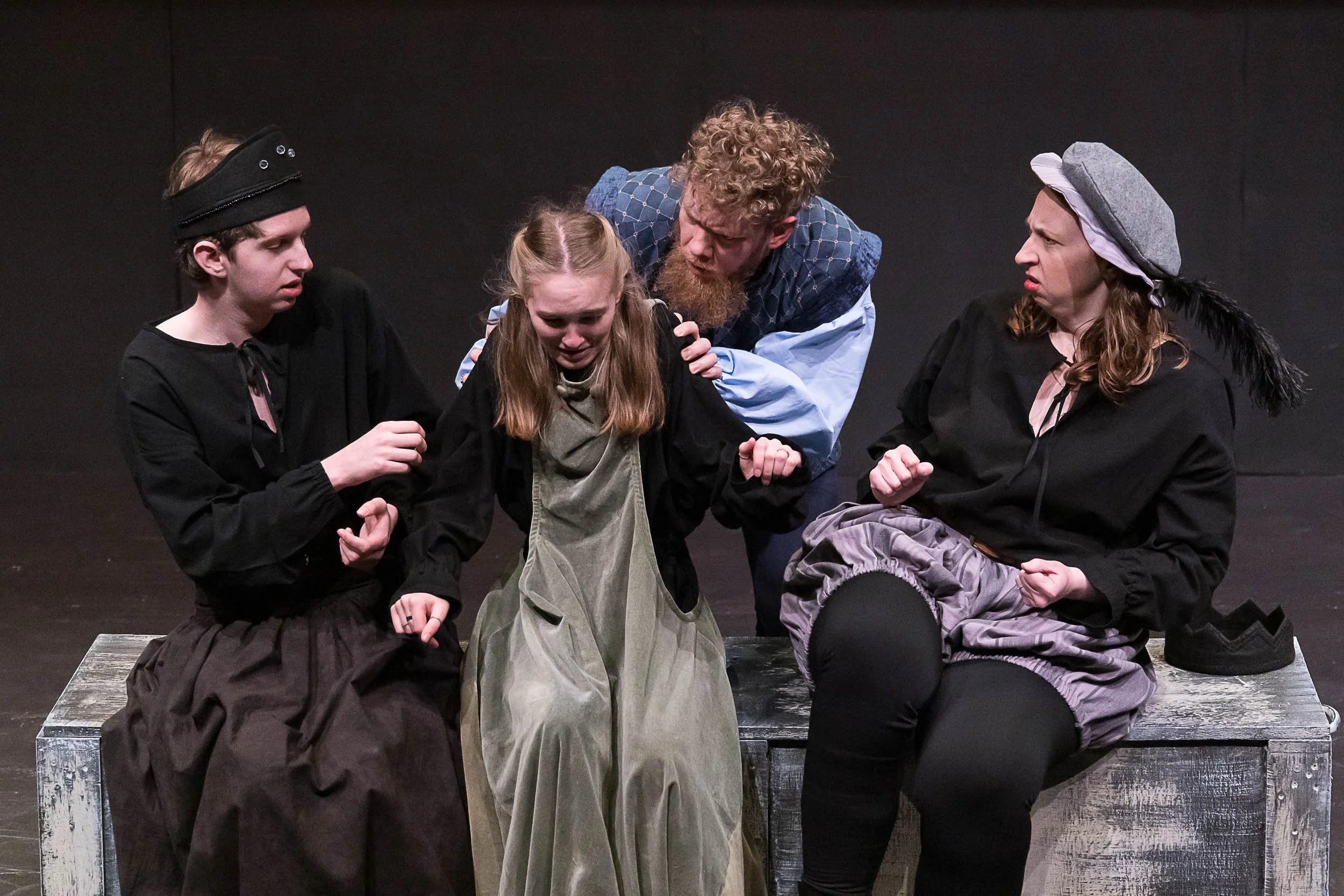 Four actors in costume on stage, seated on a wooden bench, engaged in a dramatic scene. The background is black.