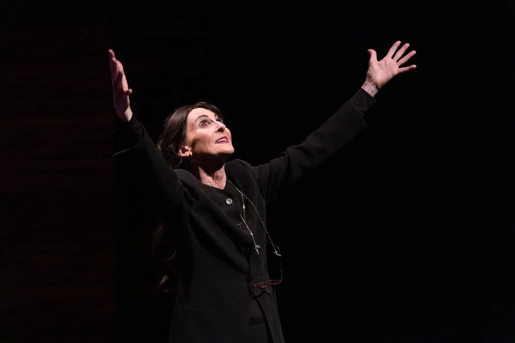 A woman in black clothing with arms raised and looking upward, standing against a dark background.