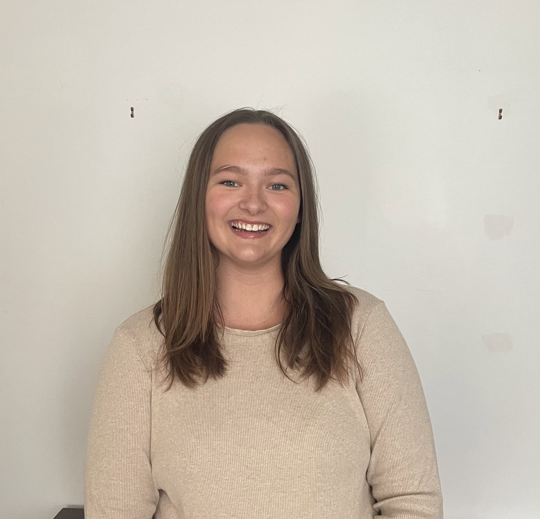 A smiling woman with long brown hair wearing a beige long-sleeve top standing against a plain white wall.