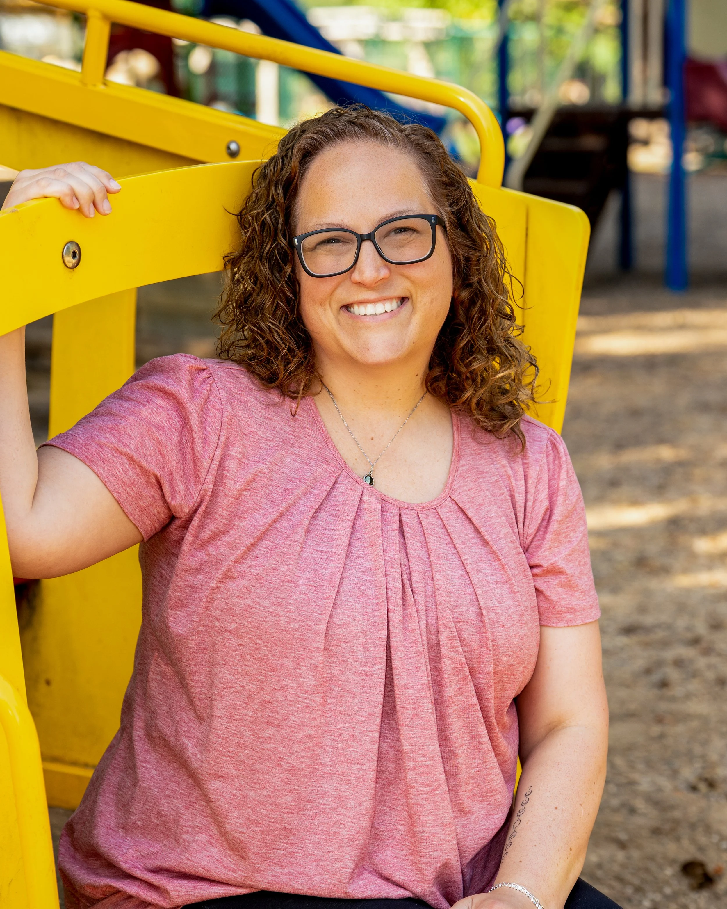 A woman with curly hair and glasses smiling, sitting outdoors near a yellow playground structure with colorful play equipment in the background.