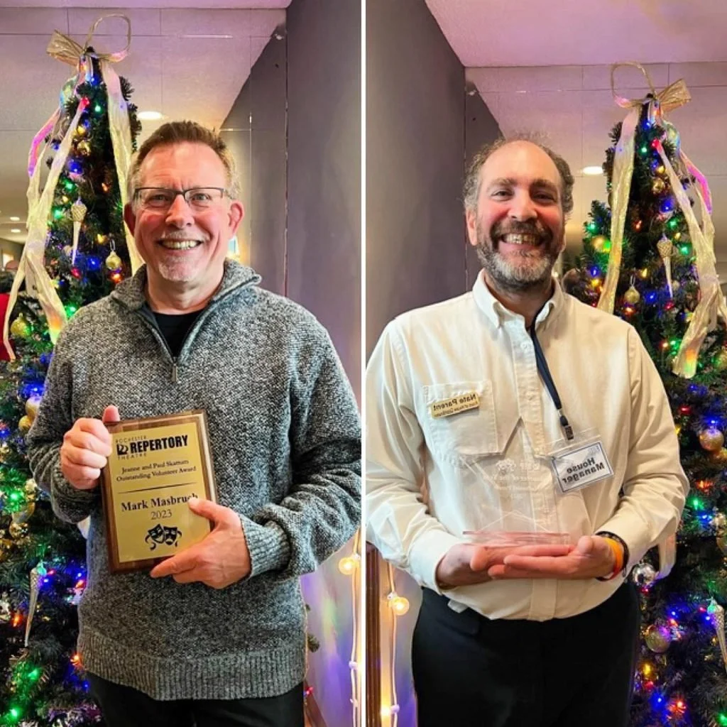 Two smiling men standing in front of decorated Christmas trees with colorful lights. The man on the left is holding a plaque and wearing glasses and a gray sweater, while the man on the right is wearing a white shirt and a name badge.