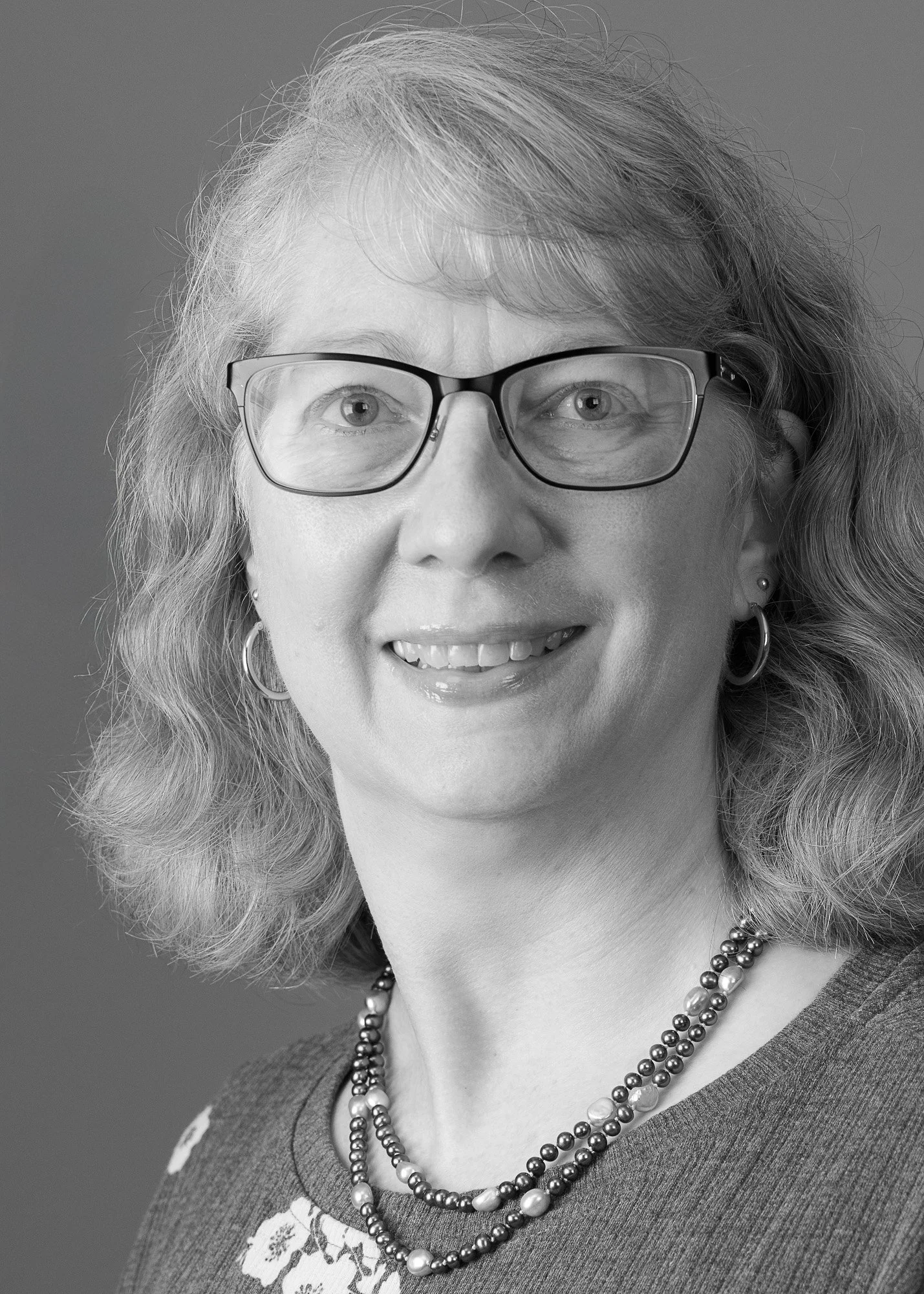 Black and white photo of a smiling woman with curly, shoulder-length hair, wearing glasses, earrings, a patterned top, and a double strand of pearls.