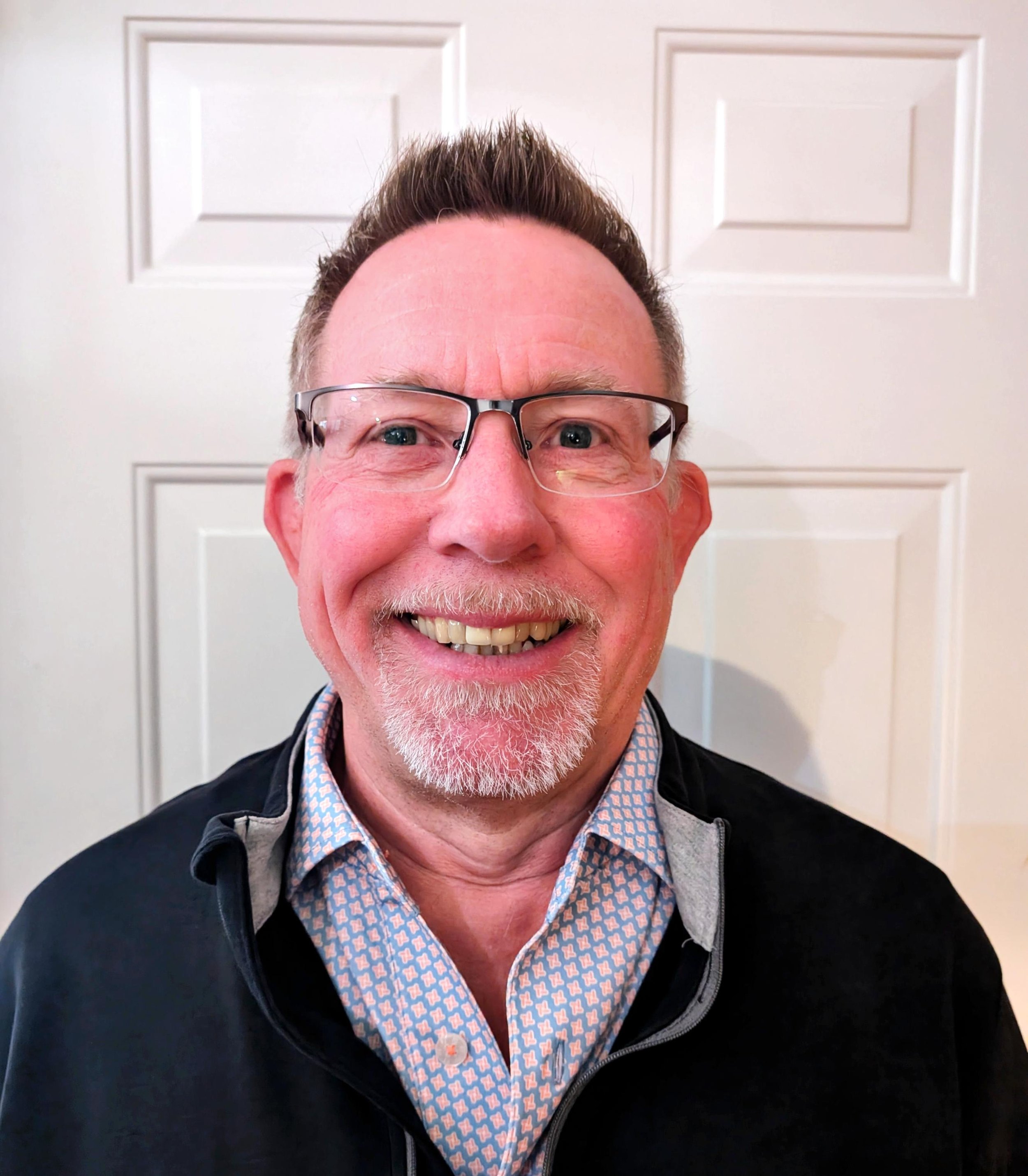 Close-up of a smiling older man with glasses, short spiked hair, and a beard, wearing a patterned shirt and black jacket, standing in front of a white paneled door.