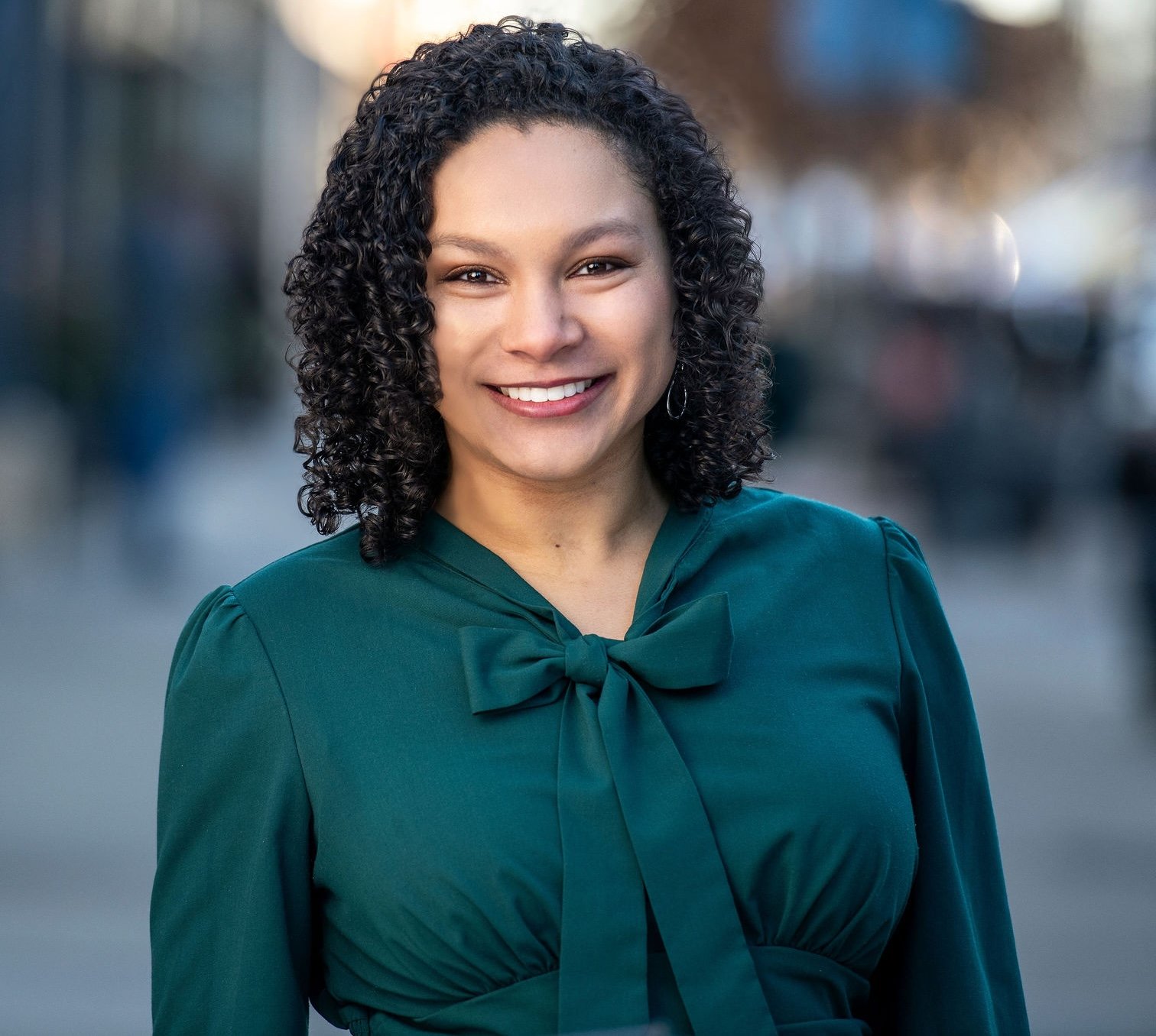 A woman with curly black hair smiling outdoors, wearing a dark green dress with a bow at the neck.