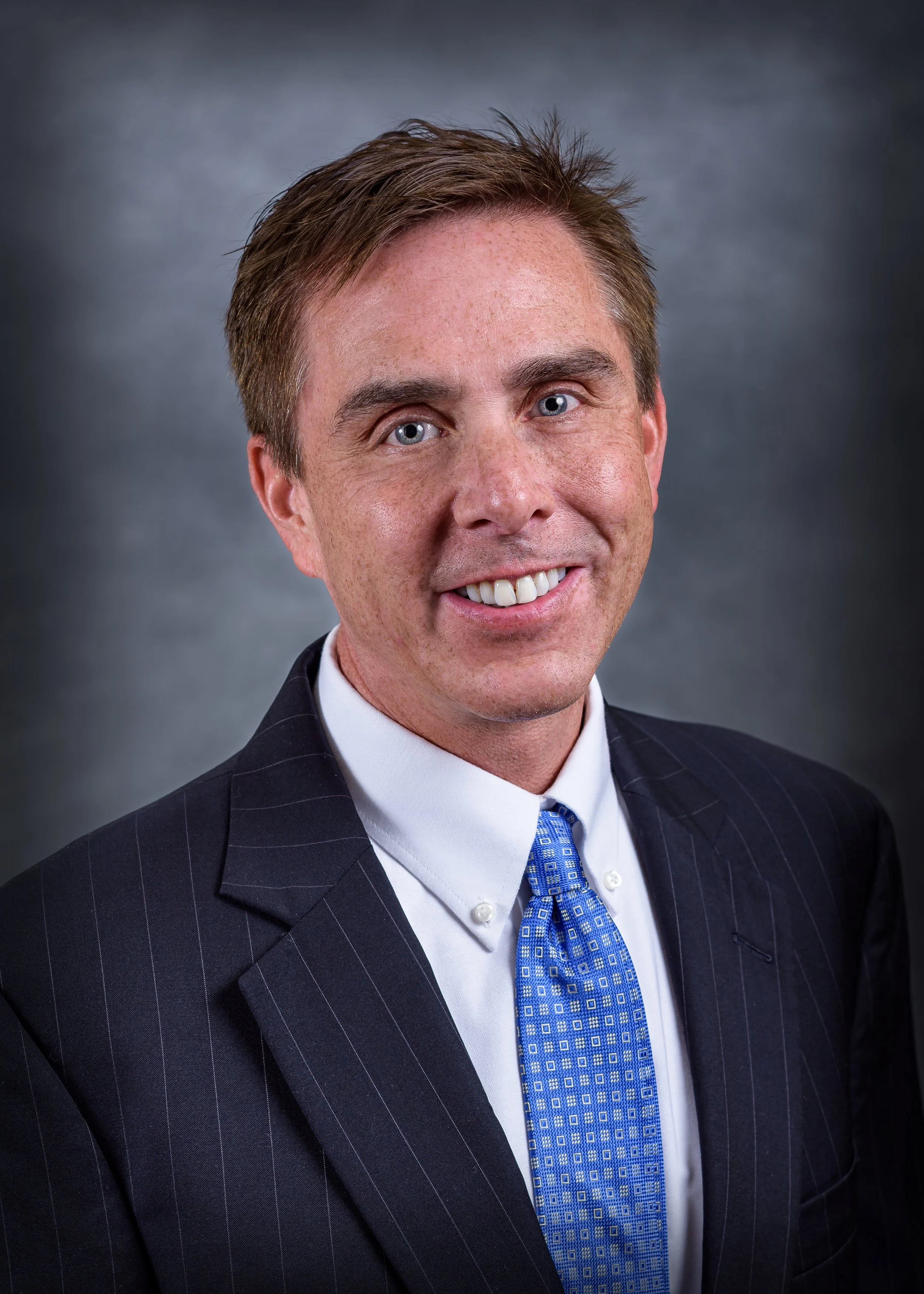 Portrait of a man in a dark pinstripe suit with a blue patterned tie, smiling, with a blurred dark background.