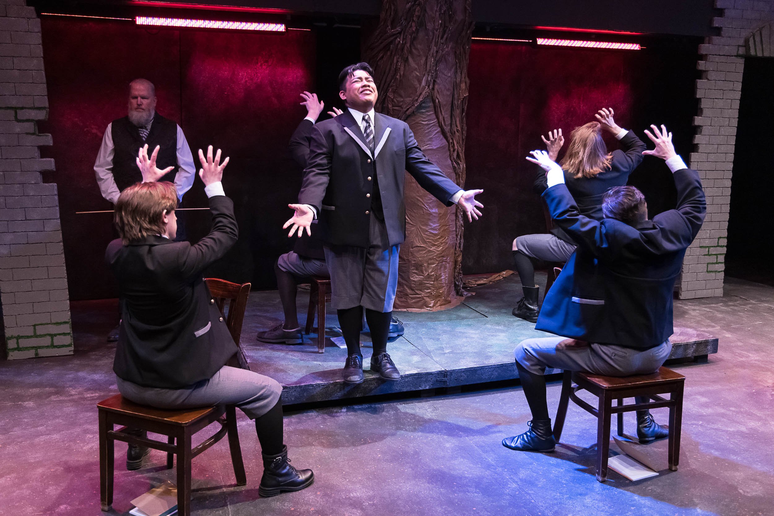 A theatrical scene with five actors, three sitting on benches and two standing, on a stage with a brick wall and a painted tree in the background. The actors are dressed in dark suits and appear to be performing, with expressive gestures and facial expressions.