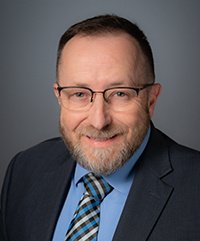 A middle-aged man with glasses, a beard, wearing a dark suit, blue shirt, and patterned blue tie, smiling against a plain gray background.