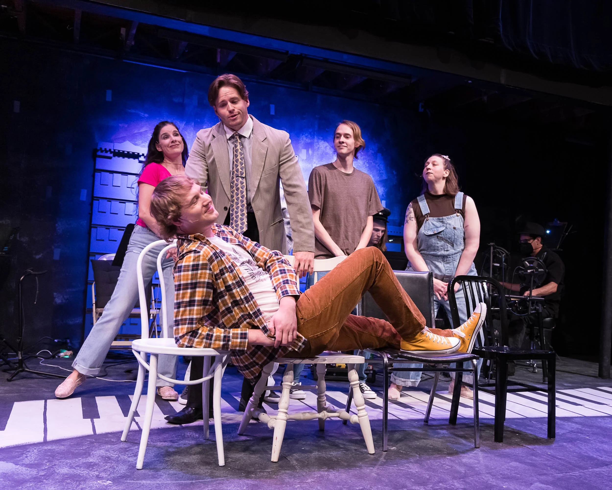 A group of six actors performing on stage, with three standing and three sitting or leaning, surrounded by chairs and theatrical equipment, under blue stage lighting.