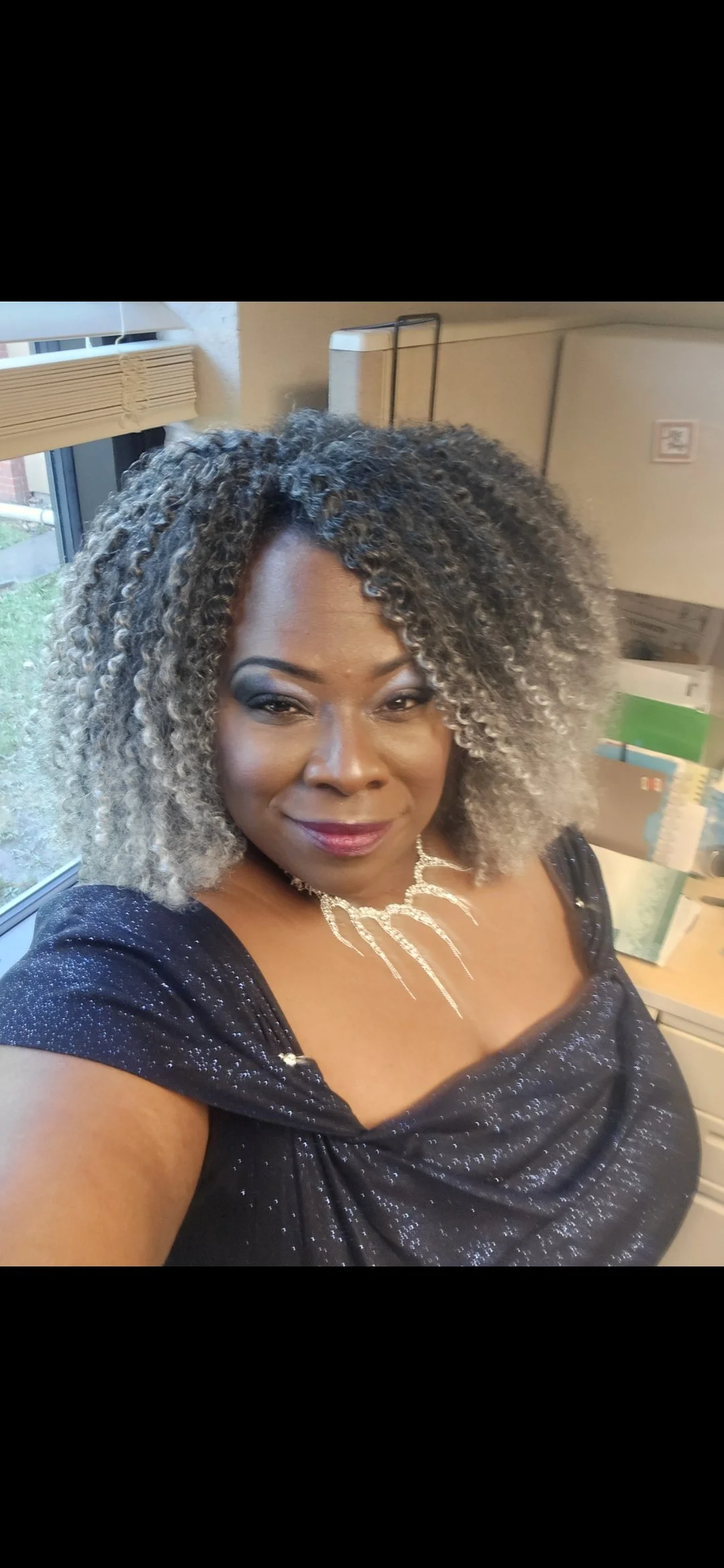 A woman with curly, gray-highlighted hair, wearing a sparkly black dress and a silver necklace, smiling indoors near a window with blinds and boxes in the background.