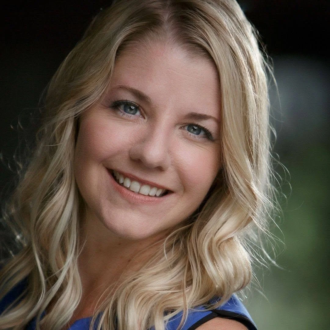 A close-up portrait of a young woman with blonde wavy hair, blue eyes, and a bright smile, wearing a blue top.