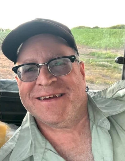 A smiling man wearing glasses, a cap, and a light-colored shirt, sitting inside a vehicle with green fields visible outside.