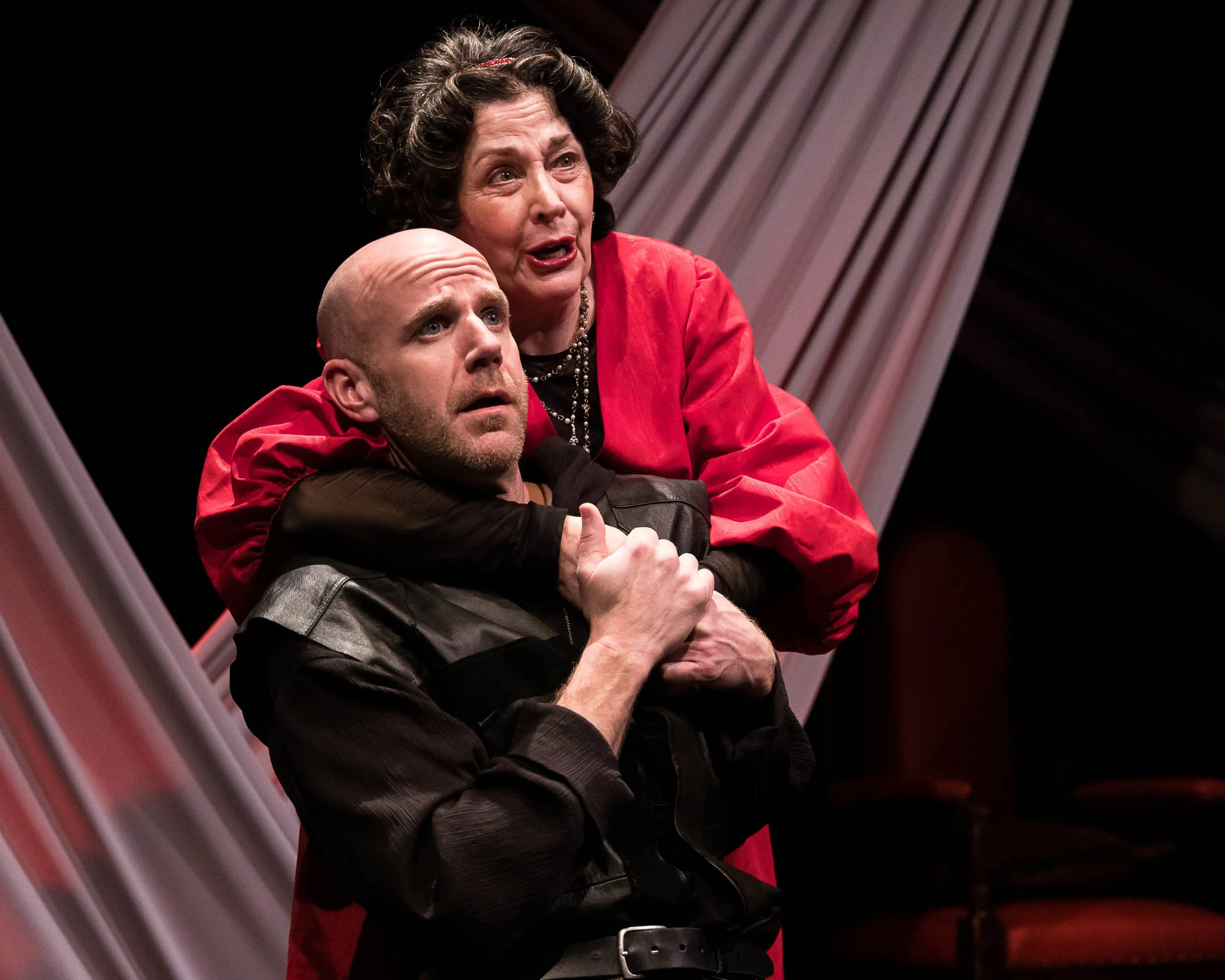 A woman with curly dark hair, wearing a red blazer and pearl necklace, is speaking passionately while embracing a bald man with a beard, dressed in a black leather jacket, behind her on a stage with draped curtains.