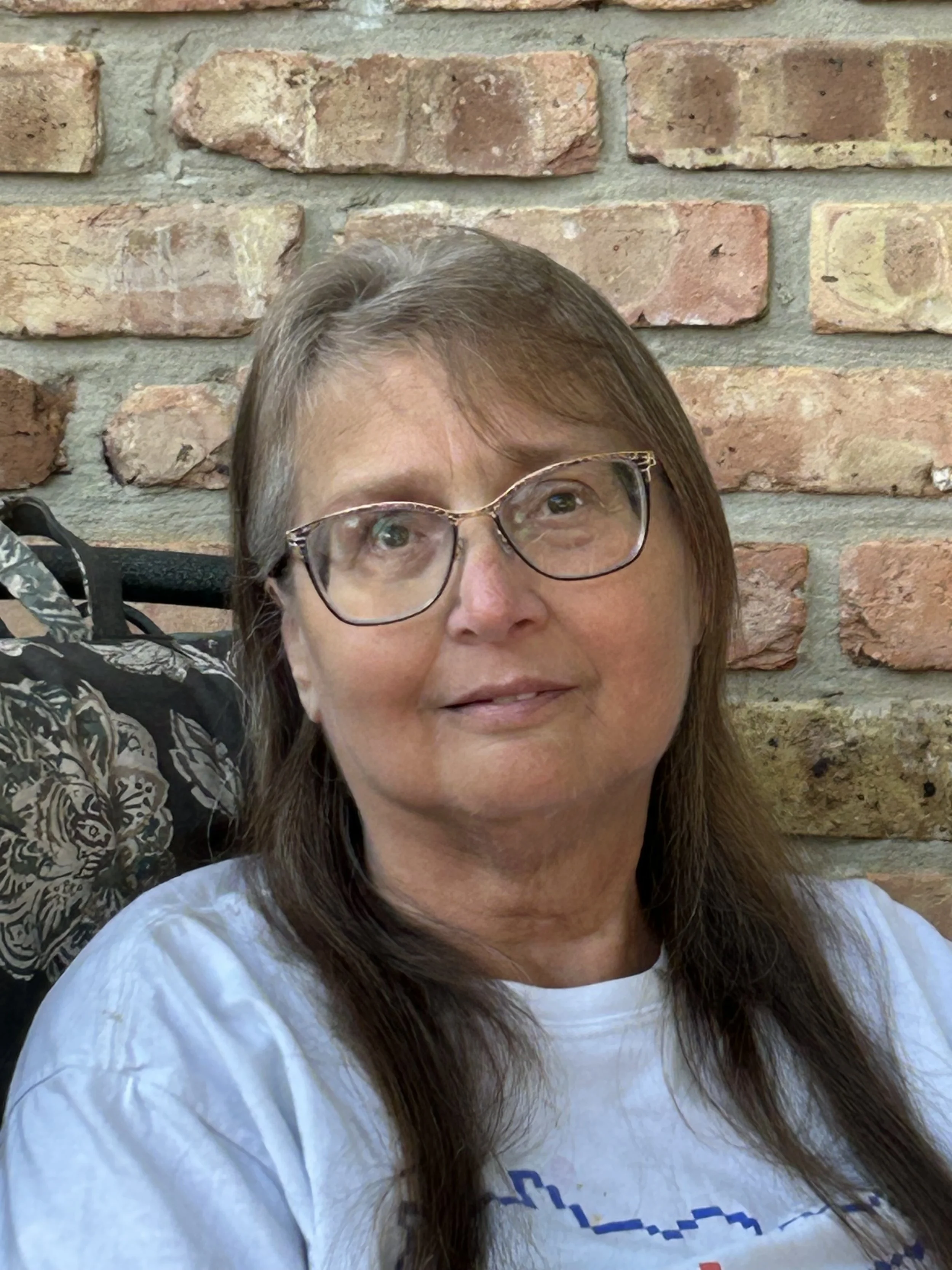 Close-up of a woman with long brown hair and glasses taking a selfie in front of a brick wall, partially visible on the lower left corner of the image.