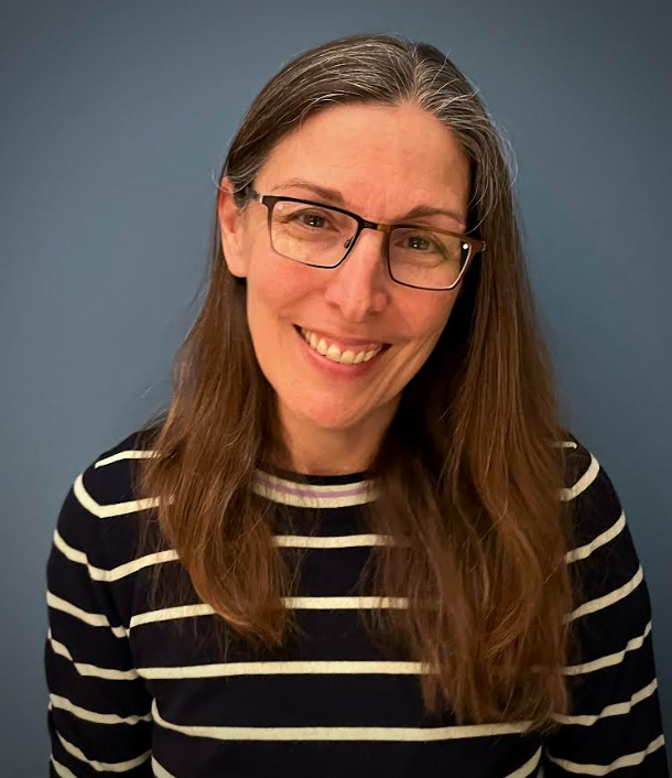 A woman with long brown hair, wearing glasses and a black and white striped shirt, smiling and standing against a blue background.