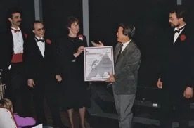 A man in a gray jacket presenting an award or framed certificate to four women and one man, all dressed in formal black tuxedos, at an indoor event.