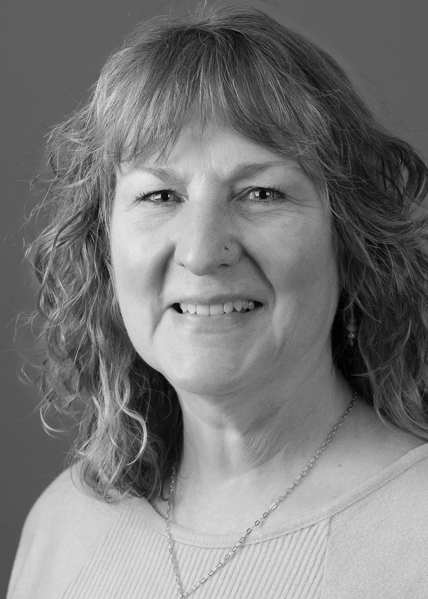 Close-up of smiling woman with curly hair and a nose piercing, wearing a necklace and a light-colored top, in black and white.