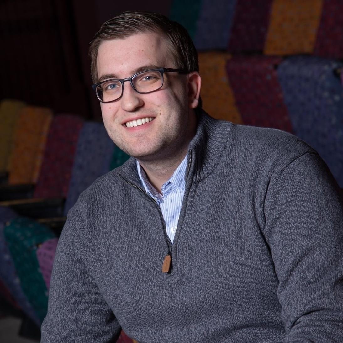 Portrait of a young man with glasses and a smile, wearing a gray zip-up sweater over a collared shirt, seated in front of colorful quilted seating.