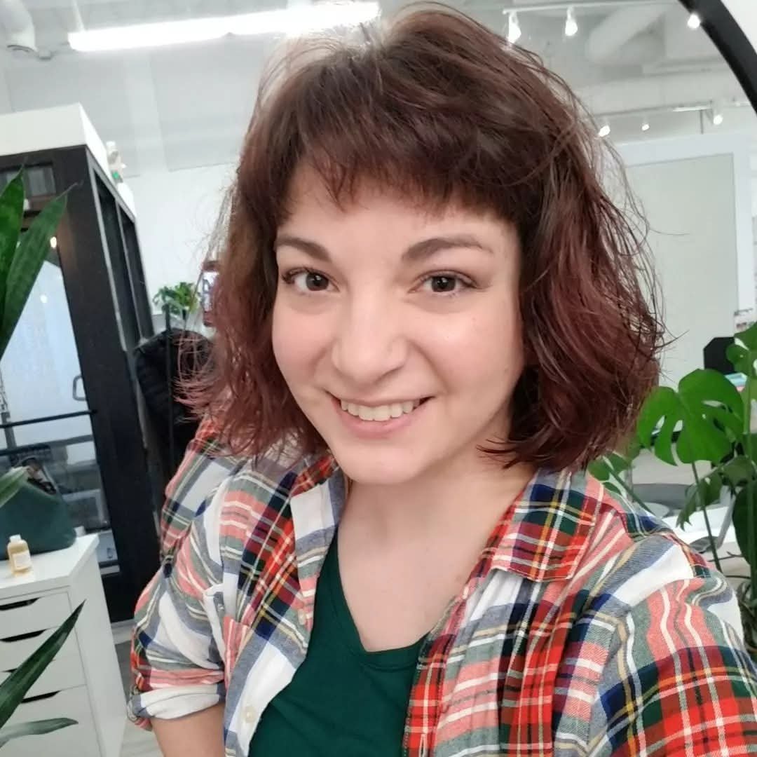 A woman with short, curly, reddish-brown hair smiling at the camera in an indoor setting with plants and modern furniture.