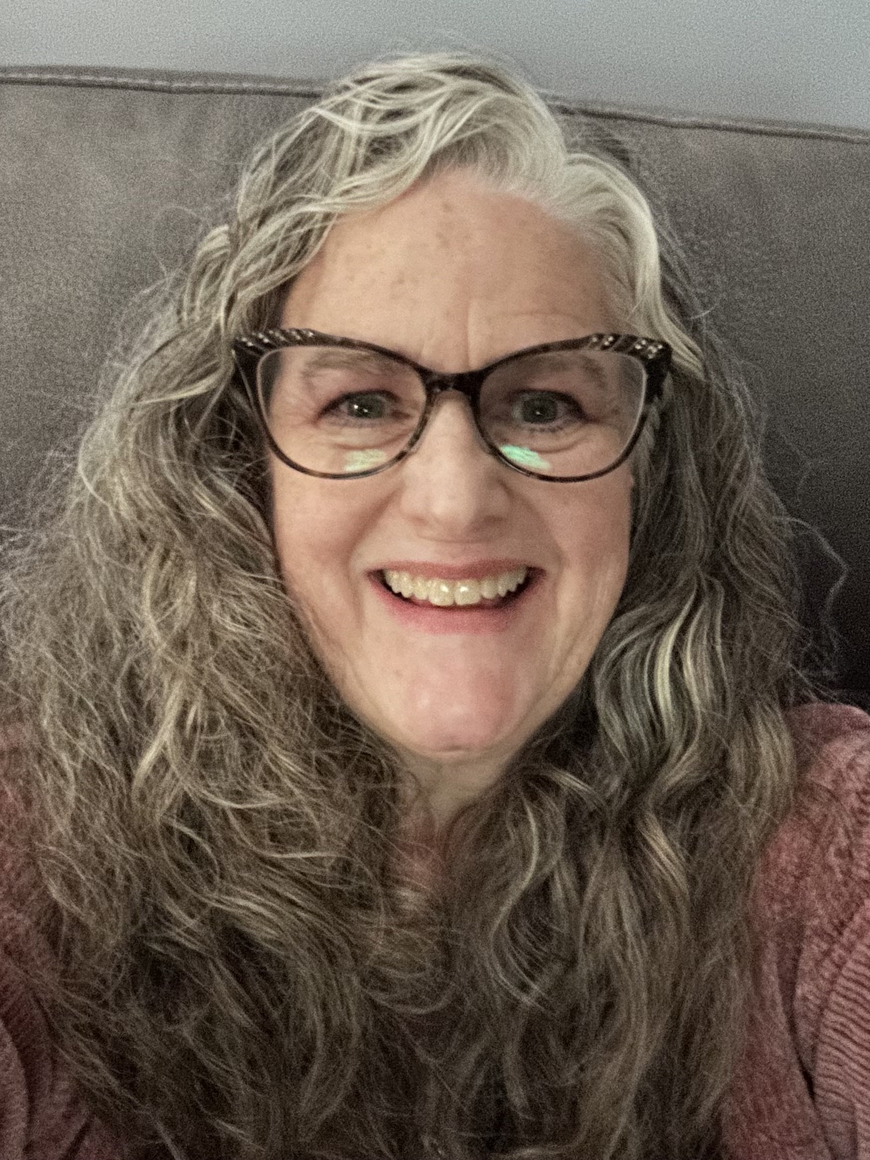 Close-up of a middle-aged woman with grey curly hair, wearing black framed glasses and smiling broadly, showing her teeth, against a grey background.