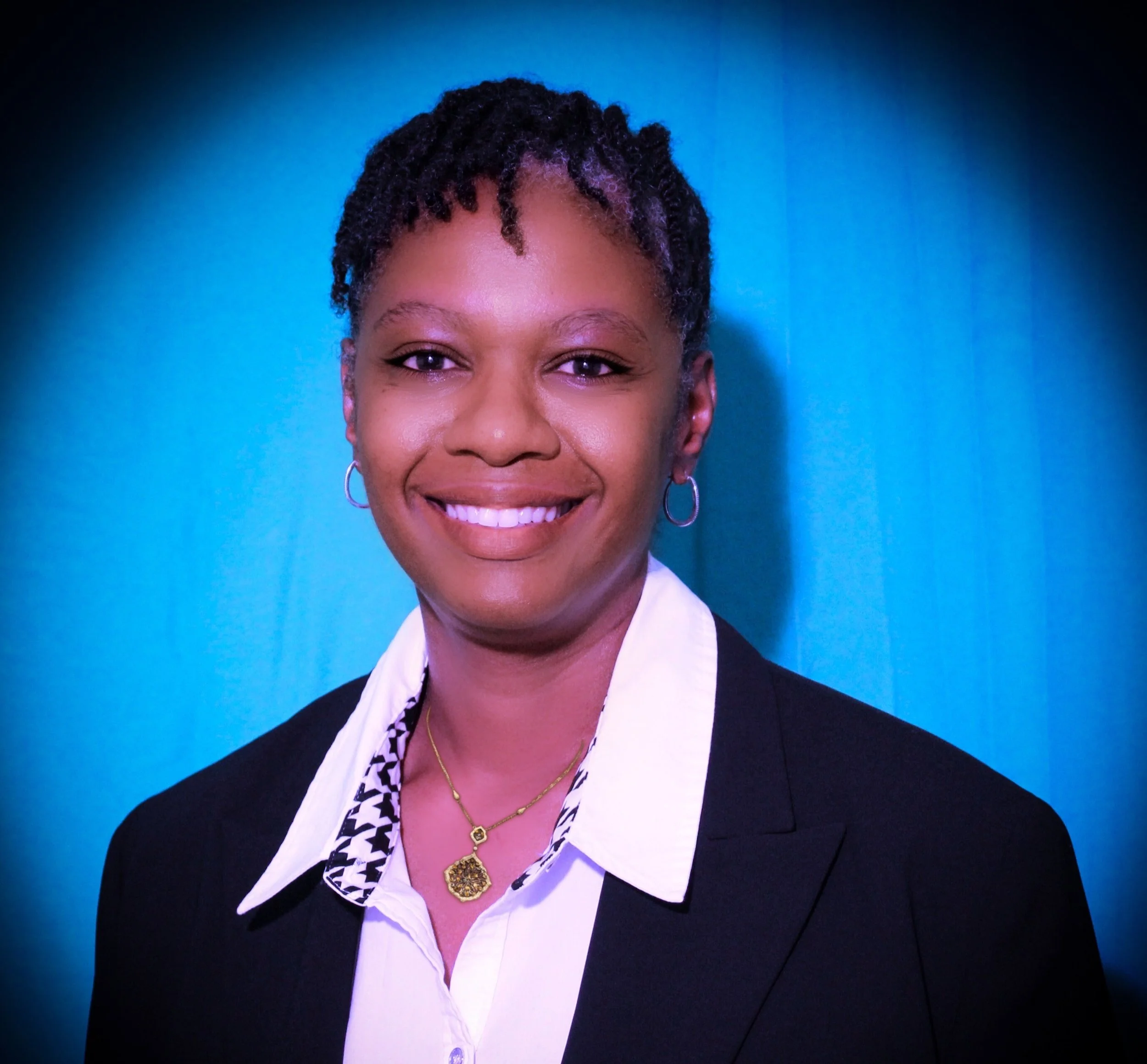 Close-up portrait of an African American woman smiling, wearing a black blazer, white shirt with a patterned collar, and jewelry, against a blue background.