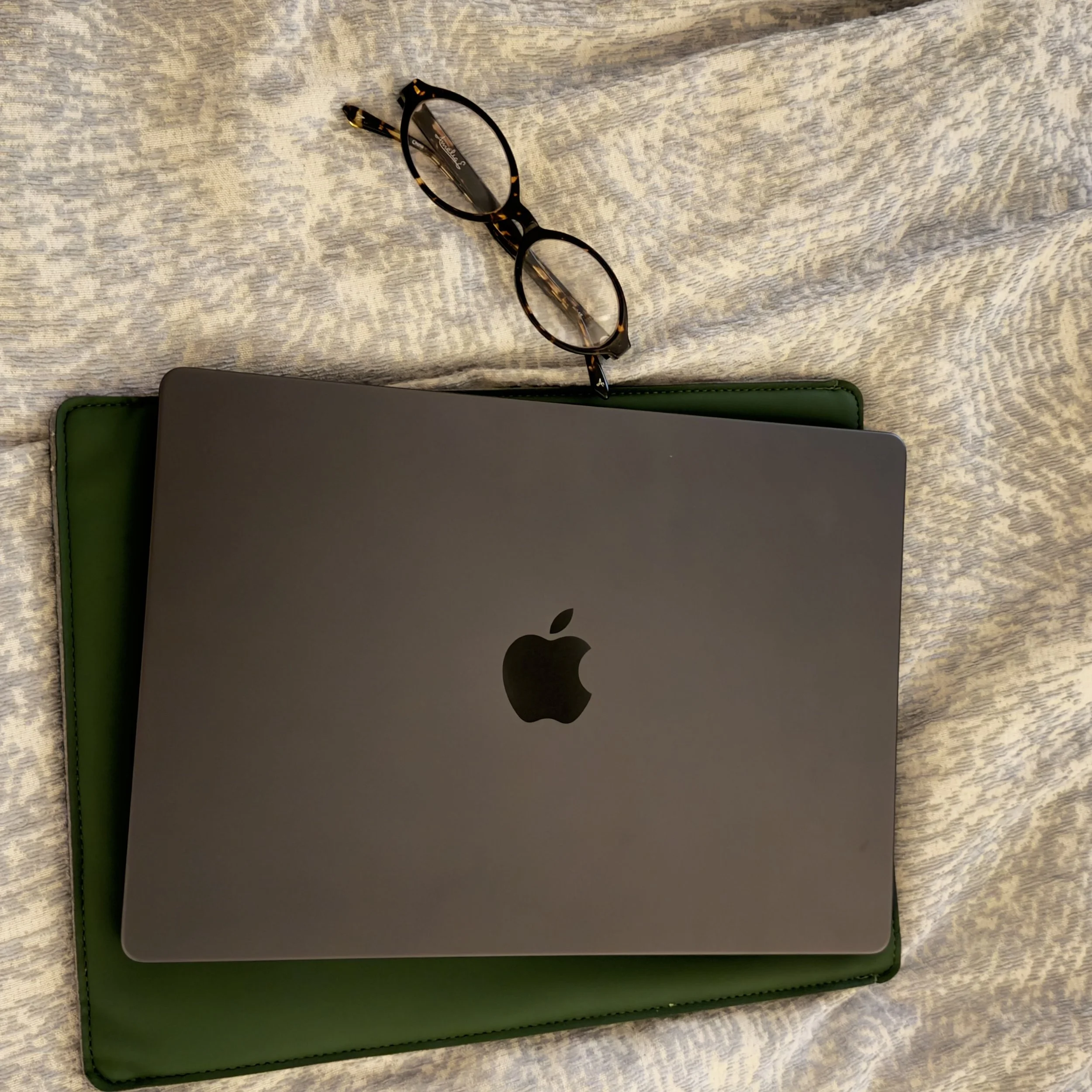 A pair of tortoiseshell eyeglasses resting on a laptop with a green case, on a textured fabric surface.