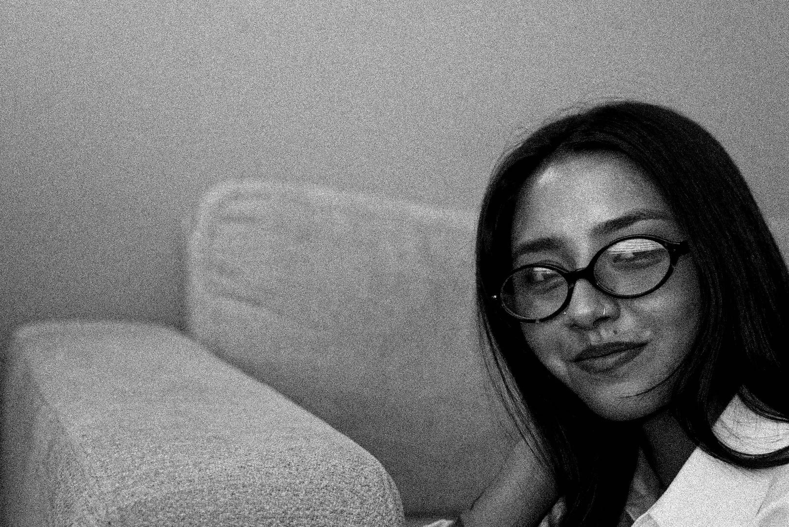 A black and white photo of a woman with long dark hair, wearing glasses, smiling softly, with a man lying beside her, partially visible, against a plain background.