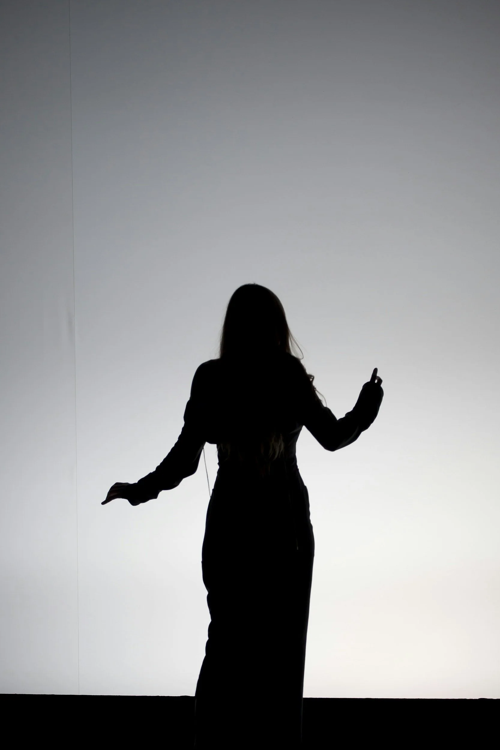 Silhouette of a woman standing in front of a light gray background, with her right hand slightly raised and her left hand pointing upward.