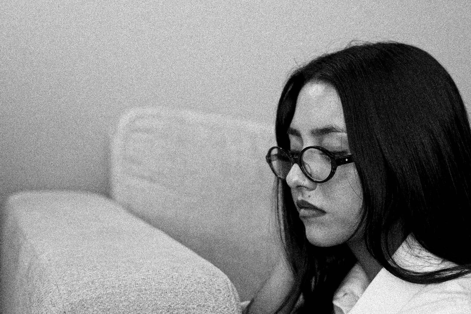 This black and white image shows a woman with long dark hair, wearing glasses, with her eyes closed, resting her face against a textured surface, possibly a pillow or cushion, in a peaceful pose.