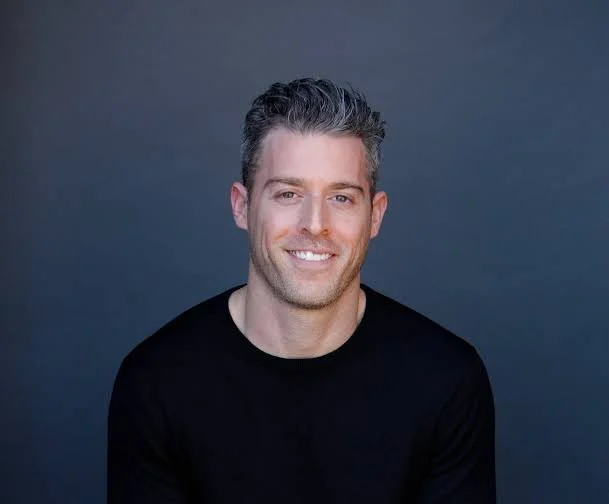 A smiling man with short, dark, slightly wavy hair wearing a black shirt against a dark gray background.