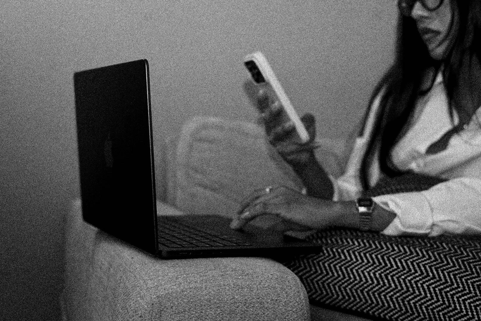 A person with long dark hair and glasses, wearing a white/striped shirt, sitting at a table, using a smartphone, with a laptop in front of them in a dimly lit room.