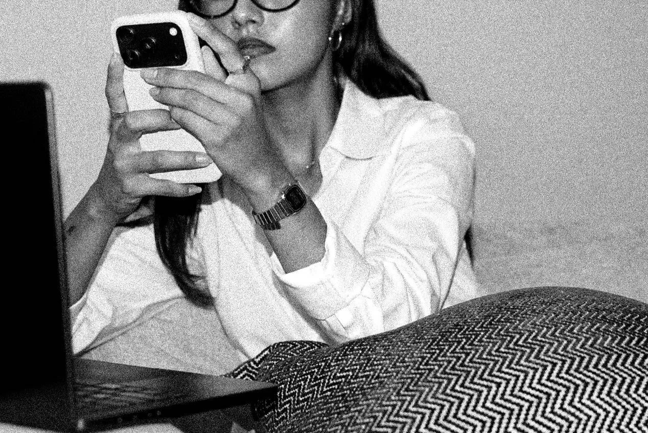 A woman wearing glasses and a watch is taking a selfie with her phone, sitting beside a laptop, in black and white.