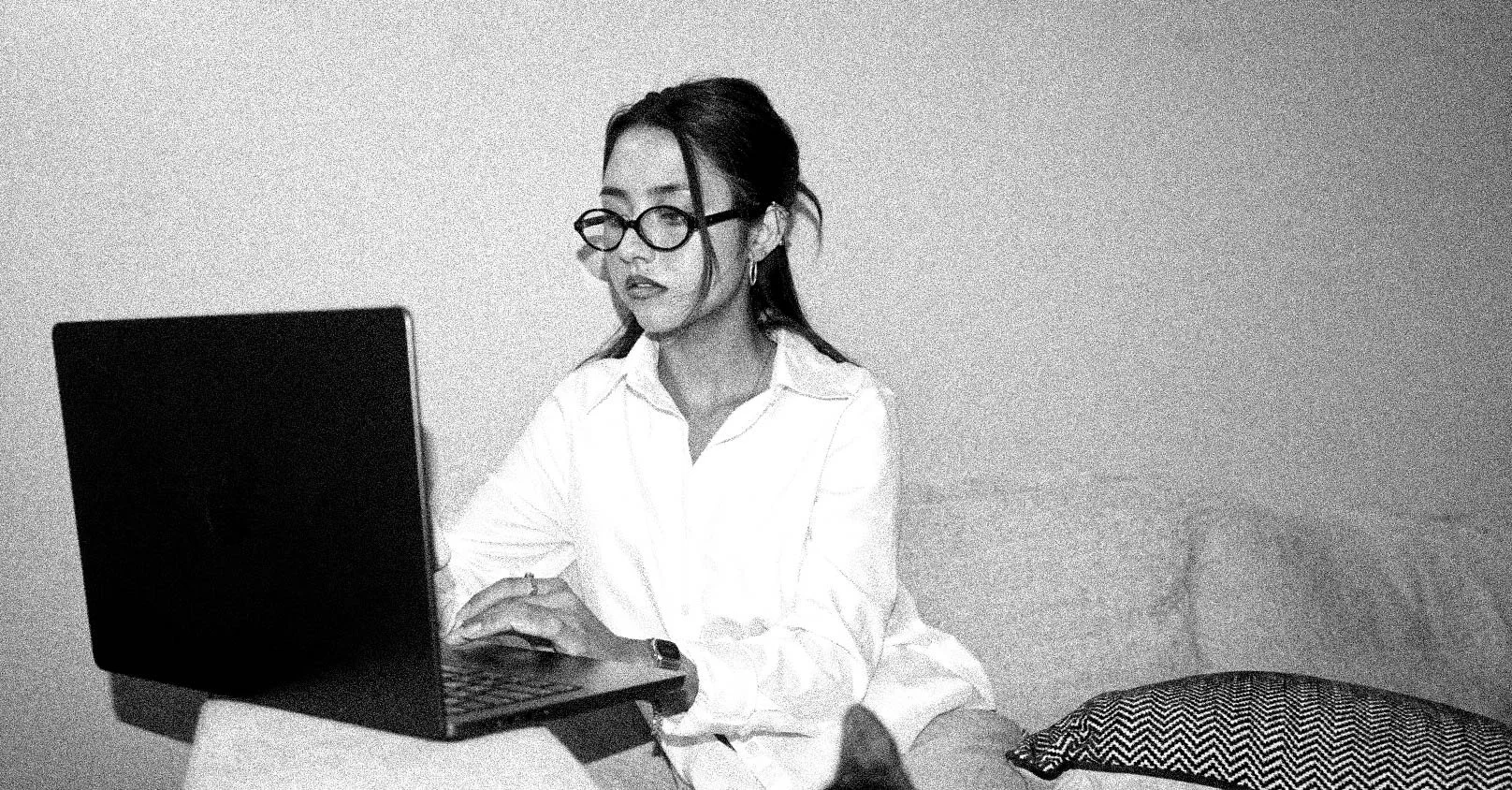 A woman with glasses and earrings, wearing a white shirt, sitting at a table working on a laptop in a room with plain walls and a patterned cushion in the background.