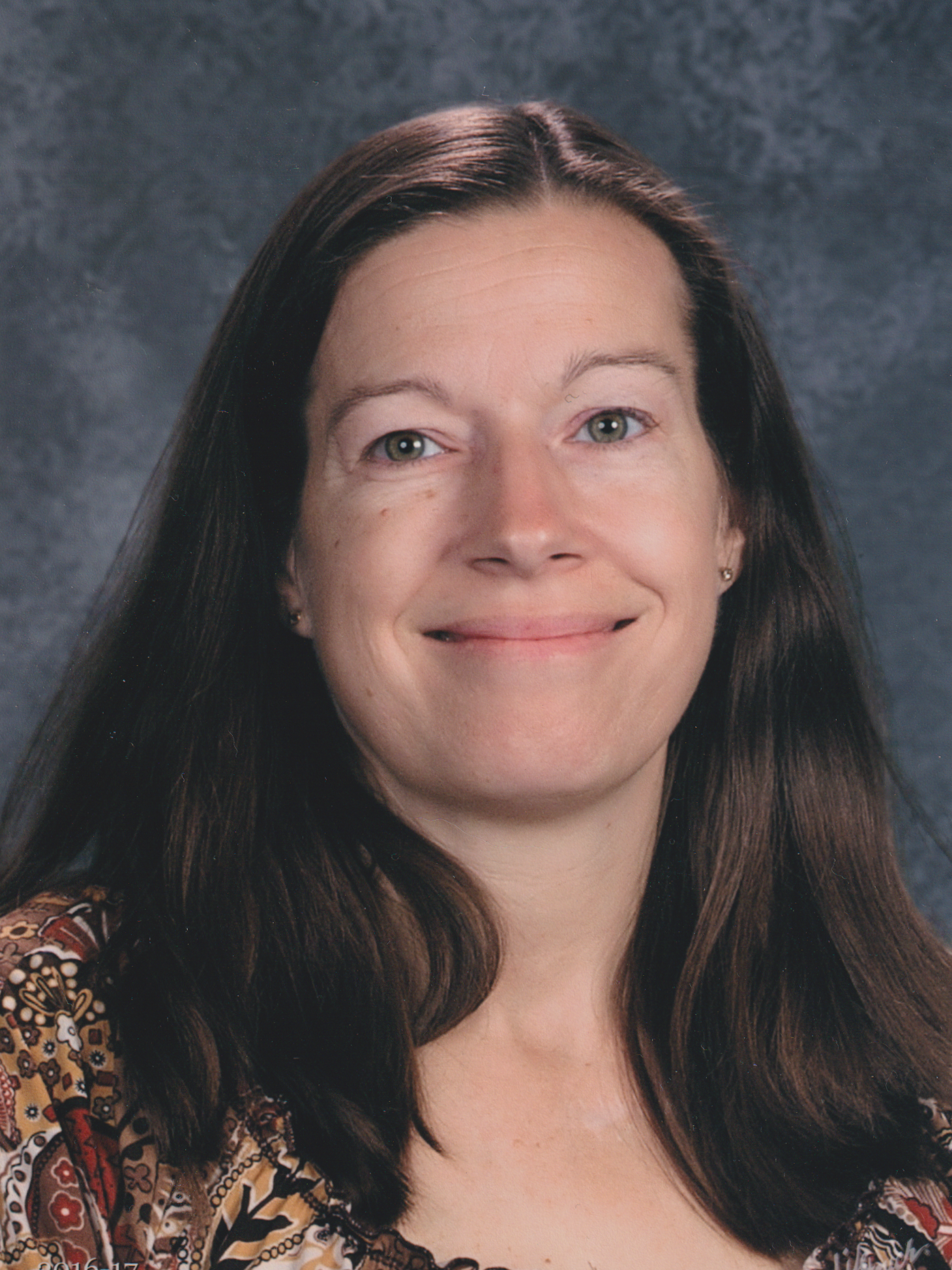 A woman with straight, dark brown hair and light skin smiling, wearing a dress with a heart, floral, and geometric pattern. She has a dark background behind her.