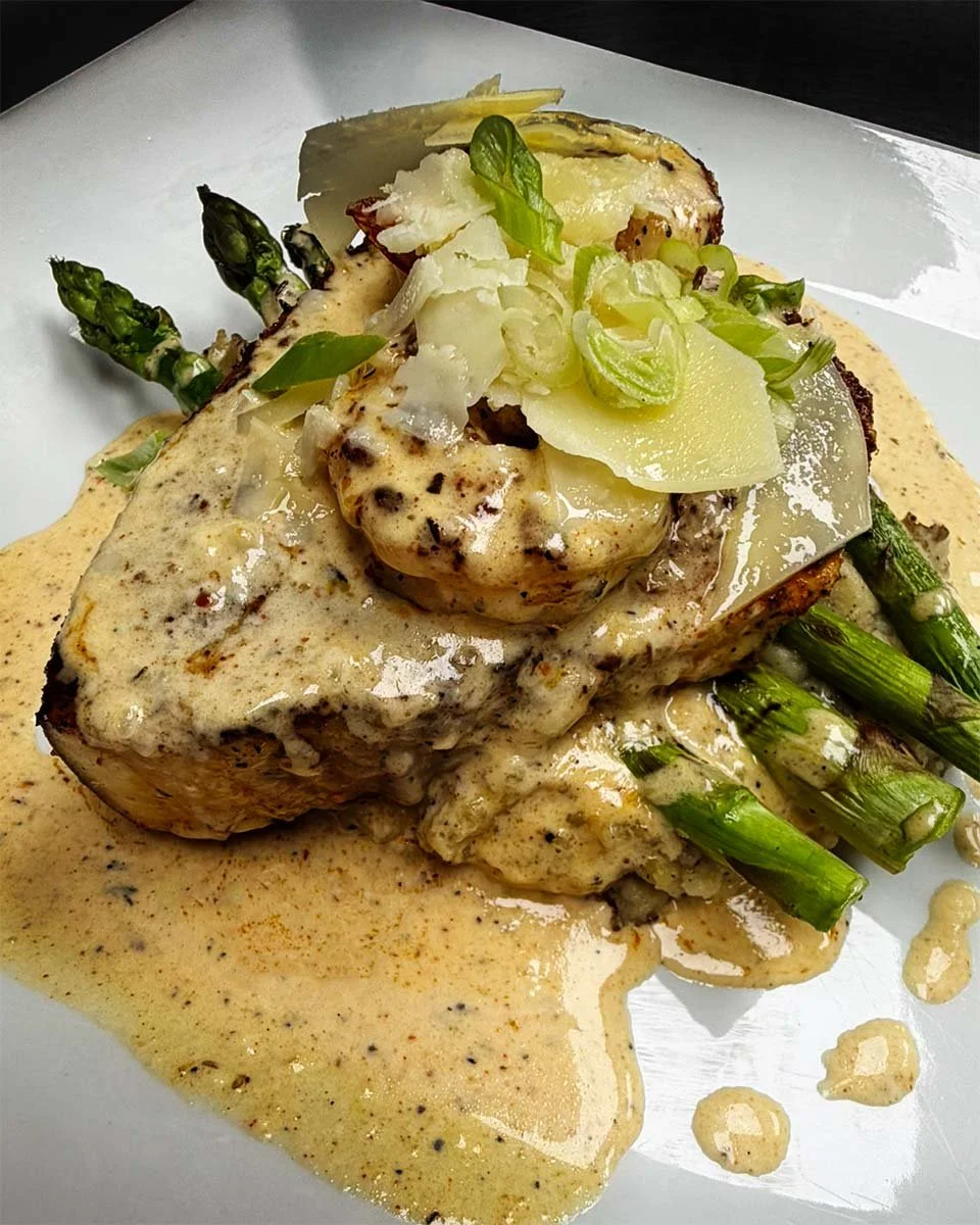 A plated dish featuring a piece of grilled chicken topped with a creamy sauce, served with grilled asparagus, topped with shaved cheese and chopped green onions, on a white plate with a drizzle of sauce around the meat.