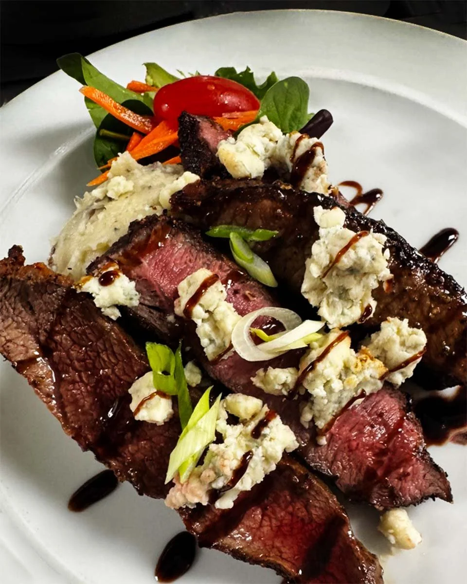 A plate with slices of grilled steak topped with blue cheese crumbles and a drizzle of balsamic glaze, served with mashed potatoes and a side salad of mixed greens, cherry tomatoes, and shredded carrots.