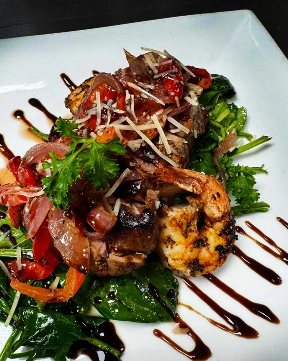 Grilled meat dish topped with shredded cheese and vegetables, served on a bed of greens with balsamic glaze on a white plate.