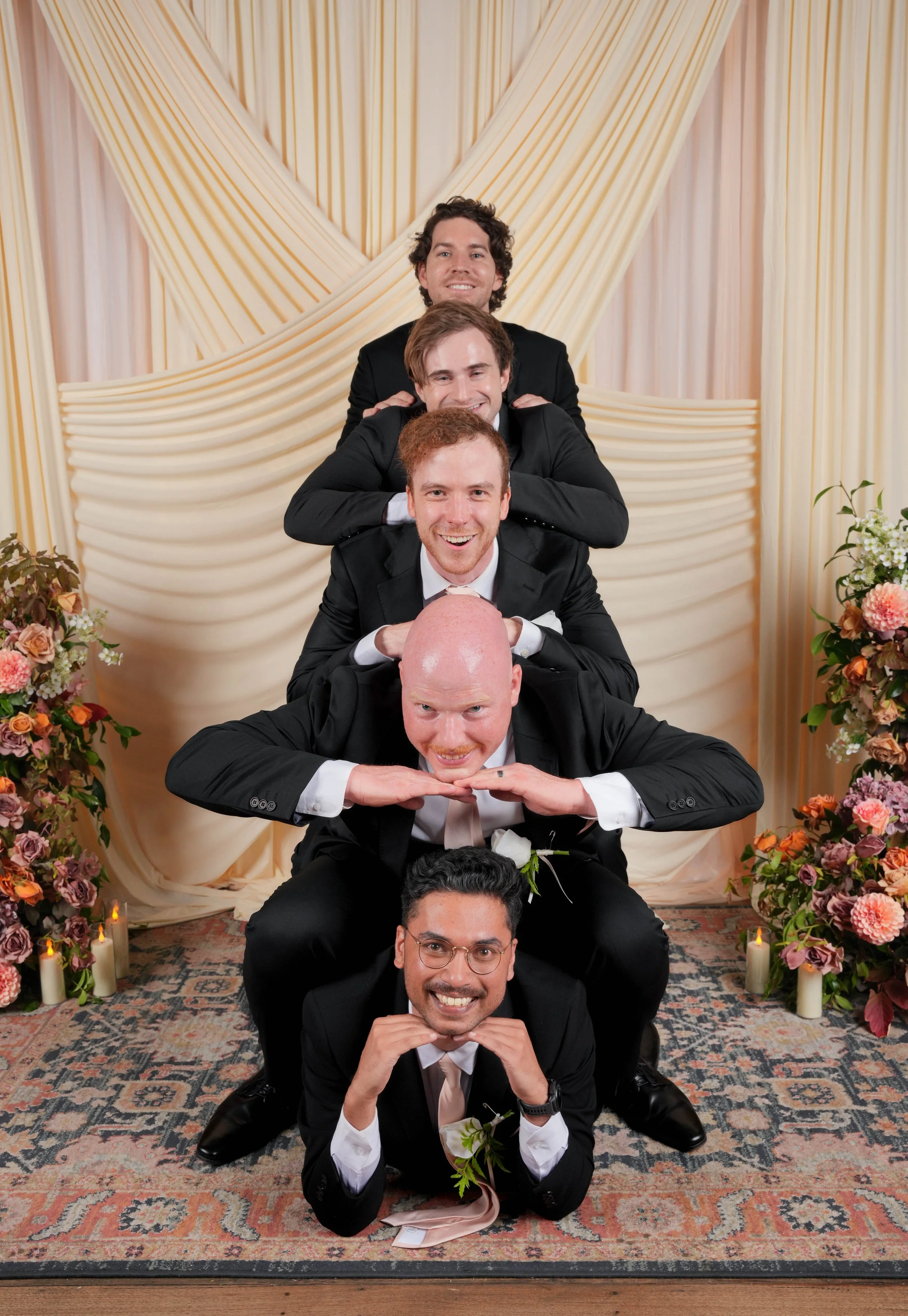 Group of six men stacking on top of each other in a pyramid pose at a wedding reception, with floral decorations and candles around them.