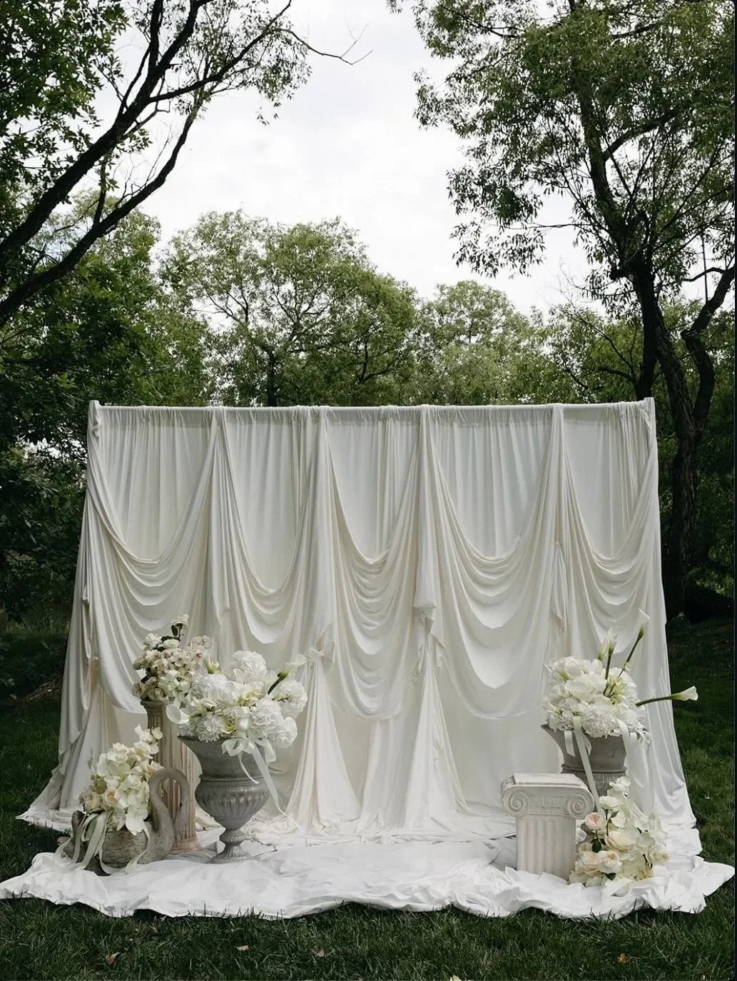 White draped backdrop with floral arrangements and small columns outdoors in a park or garden.