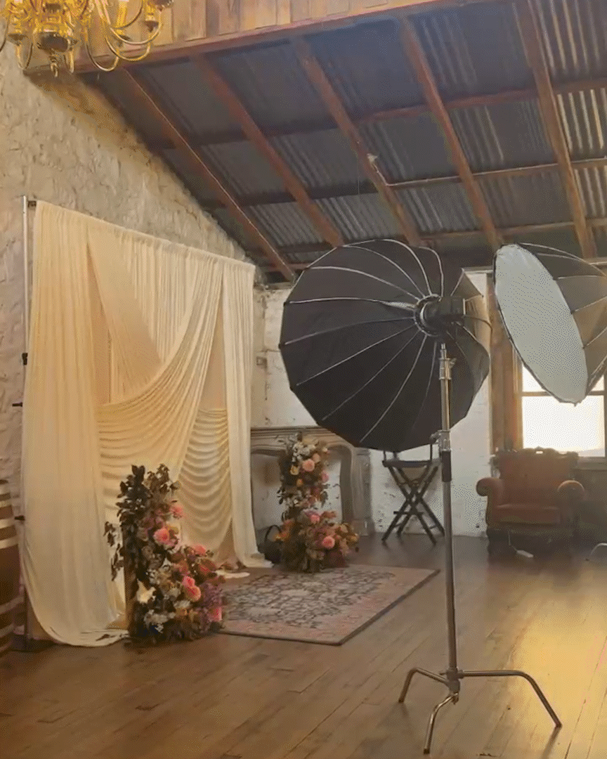 Photo of a photography or video studio setup with professional lighting, a fabric backdrop, floral arrangements, a chair, and a vintage sofa in a rustic room with a corrugated metal ceiling.