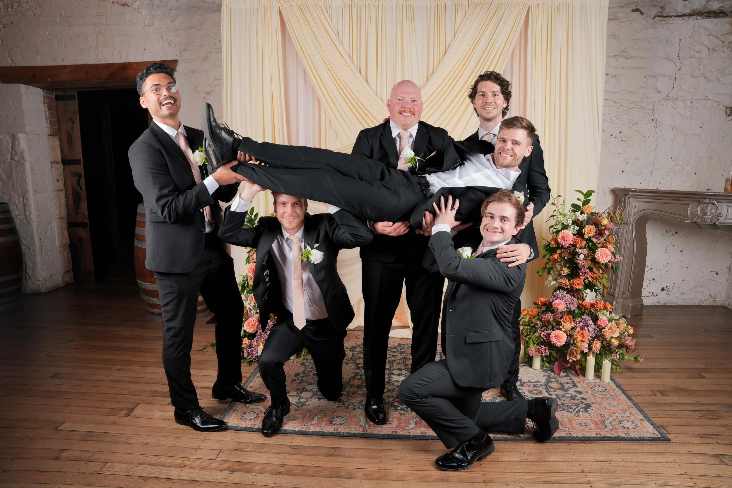 A group of six men in formal suits holding a man horizontally in a wedding or celebration setting with floral decorations and a backdrop with light-colored drapes.