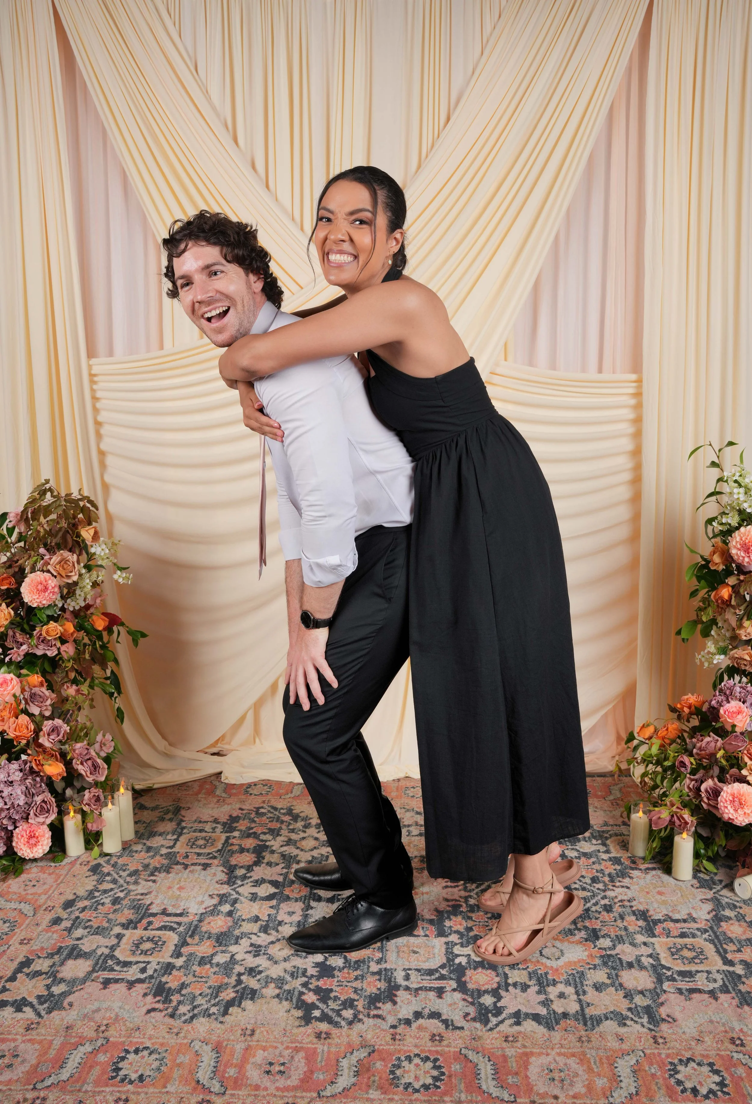 A joyful couple is at a wedding reception, with the woman giving the man a piggyback ride. They are smiling and making playful expressions. The background features a beige and cream draped backdrop, with floral arrangements and lit candles on the sides. The man is wearing a white shirt, black pants, and black shoes, while the woman is wearing a black dress, beige sandals, and earrings. They are standing on a patterned rug.